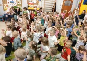 Przedszkolaki siedzą na dywanie i machają do rytmu.
