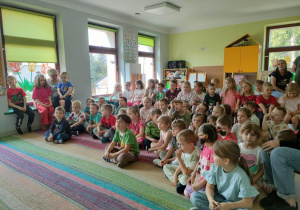 Starsze dzieci siedzą na dywanie i uważnie słuchają przybyłych Gości.
