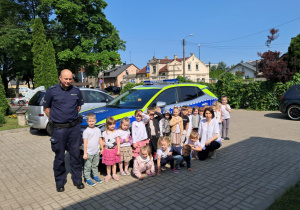 Zdjęcie dzieci z grupy "Żabek" wraz z nauczycielką oraz Funkcjonariuszem Policji na tle radiowozu.