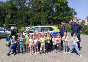 Zdjęcie dzieci z grupy "Motylków" wraz z nauczycielką oraz Funkcjonariuszem Policji na tle radiowozu.