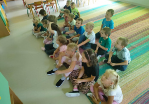 Dzieci siedzą na dywanie i z uwagą oglądają krótką bajkę edukacyjną, dotyczącą zdrowego odżywiania.