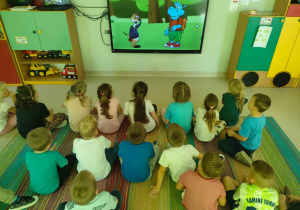 Dzieci siedzą na dywanie i z uwagą oglądają krótką bajkę edukacyjną, dotyczącą zdrowego odżywiania.