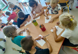 Kilkoro dzieci przy stoliku rysuje ołówkiem po śladzie szablon sportowej koszulki.