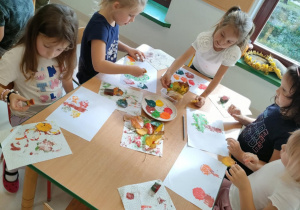 Dzieci przy stoliku malują jesień, z wykorzystaniem naturalnych, nietypowych pędzli, jakim są liście.