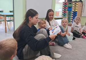 Jedna z uczennic czyta dzieciom z grupy "Żabek" bajkę "Kicia Kocia". Dzieci siedzą na dywanie i słuchają treści.