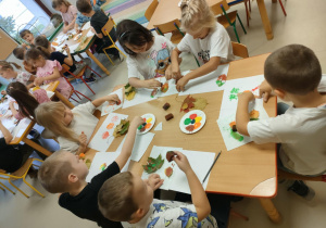 Kilkoro dzieci przy stoliku stempluje kartkę pomalowanymi liśćmi.