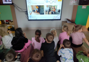 Panie prowadzące spotkanie online, prezentują dzieciom umiejętności dwóch psów: Holki i Morki. Dzieci siedzą na dywanie, jednocześnie jedząc jabłuszko.