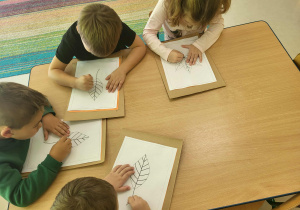 Kilkoro dzieci przy stole nakłuwa szablon jesiennych liści.
