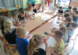 Dzieci siedza przy połączonych stołach i malują swoje dłonie, przy wykorzystaniu farb w jesiennych kolorach i gąbeczek.