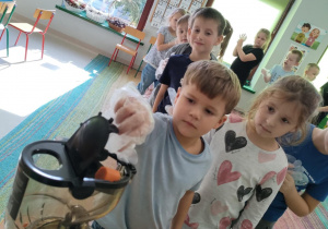 Dzieci kolejno, w rękawiczkach wrzucają kawałki jabłka/marchewki do wyciskarki wolnobrotowej.