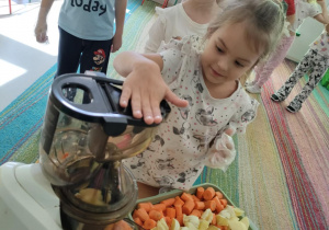 Dzieci kolejno, w rękawiczkach wrzucają kawałki jabłka/marchewki do wyciskarki wolnobrotowej.