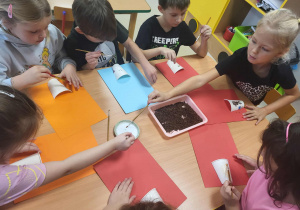 Dzieci za pomocą pędzelków, nakładają klej na kubeczek, przyczepiony do kolorowej kartki.