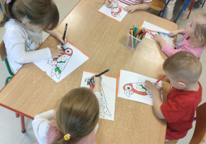 Kilkoro dzieci przy stoliczku koloruje szablon papugi zgodnie z podanymi kolorami.