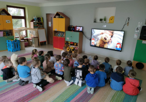 Połączone grupy maluszków i starszaków oglądają film edukacyjny o Dniu Pluszowego Misia na ekranie multimedialnym.