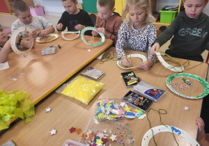 Dzieci siedzą przy połączonych stołach i przewlekają przez dziurki zieloną wełnę.