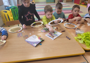 Dzieci siedzą przy połączonych stołach i przewlekają przez dziurki zieloną wełnę.