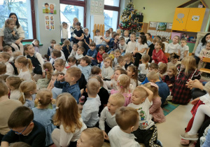 Przedszkolaki siedzą na dywanie podczas wigilijnego spotkania z Księdzem.