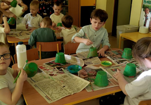 "Motylki" malują zieloną farbą małą doniczkę.