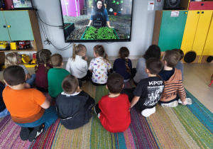 Dzieci oglądają film edukacyjny o rukoli na ekranie multimedialnym.