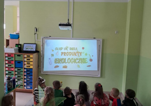 Dzieci z grupy "Żabek" oglądają film edukacyjny pt. "Skąd się biorą produkty ekologiczne?" na tablicy interaktywnej.