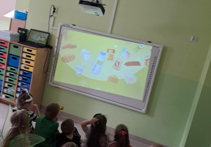 Dzieci z grupy "Żabek" oglądają film edukacyjny pt. "Skąd się biorą produkty ekologiczne?" na tablicy interaktywnej.