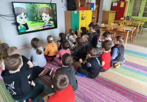 "Pszczółki" oglądają film edukacyjny pt. "Skąd się biorą produkty ekologiczne?" na ekranie multimedialnym.