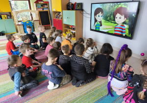 "Pszczółki" oglądają film edukacyjny pt. "Skąd się biorą produkty ekologiczne?" na ekranie multimedialnym.