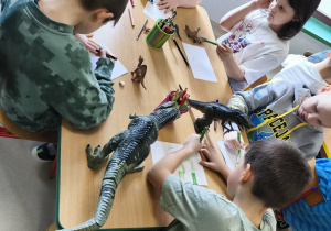 Kilkoro dzieci przy stoliku samodzielnie rysuje swojego dinozaura.