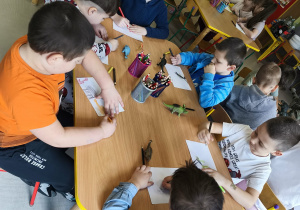 Kilkoro dzieci przy stoliku samodzielnie rysuje swojego dinozaura.