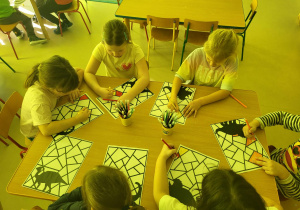 Kilkoro dzieci przy stoliku koloruje mazakami przygotowane wcześniej karty z dinozaurami.