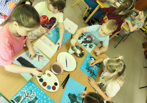 Kilkoro dzieci wraz z nauczycielką przy stole tworzy pracę plastyczną "Las zimą" z wykorzystaniem niebieskich kartek, farb i pędzelków.