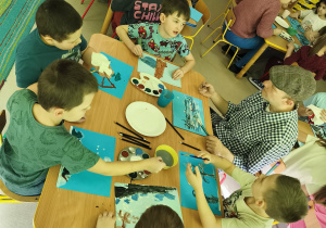 Kilkoro dzieci wraz z p. Pawłem Jaszczakiem przy stole tworzy pracę plastyczną "Las zimą" z wykorzystaniem niebieskich kartek, farb i pędzelków.