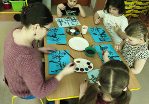 Kilkoro dzieci wraz z p. Joanną Jaszczak przy stole tworzy pracę plastyczną "Las zimą" z wykorzystaniem niebieskich kartek, farb i pędzelków.