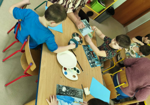 Kilkoro dzieci wraz z p. Dyrektor przy stole tworzy pracę plastyczną "Las zimą" z wykorzystaniem niebieskich kartek, farb i pędzelków.