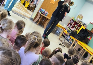 Pan Jacek gra na trąbce, a dzieci siedząc na dywanie słuchają melodii.