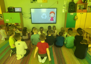 Dzieci siedzą na dywanie i oglądają bajkę edukacyjną pt. "Recykling" na ekranie multimedialnym.