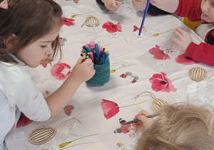 Kilkoro dzieci z najmłodszej grupy przy stoliku koloruje mazakami drewniane pisanki.