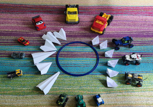 Zabawkowe i papierowe pojazdy ułożone na środku dywanu.