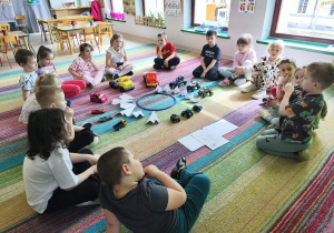 Dzieci siedzą w kręgu na dywanie, podczas rozmowy na temat transportu różnorodnych produktów.