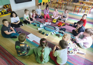 Dzieci siedzą w kręgu na dywanie, podczas rozmowy na temat transportu różnorodnych produktów.