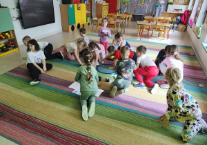 Przedszkolaki przewożą na swoich pojazdach obrazki różnych produktów na środek dywanu.