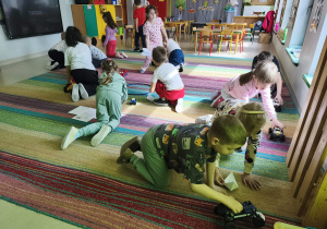 Przedszkolaki przewożą na swoich pojazdach obrazki różnych produktów na środek dywanu.