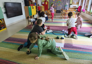 Przedszkolaki przewożą na swoich pojazdach obrazki różnych produktów na środek dywanu.