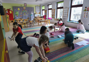 Przedszkolaki przewożą na swoich pojazdach obrazki różnych produktów na środek dywanu.