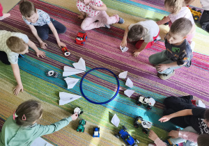 Dzieci parkują swoje, zabawkowe/papierowe pojazdy wokół obręczy.