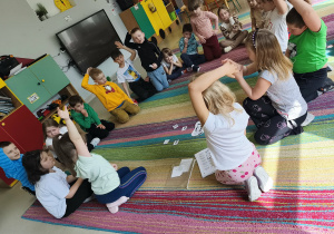 Dzieci siedzą na dywanie i podają zdrowe zamienniki dla bohaterów historyjki(Asi i Karola), którzy zbyt często sięgali po słodkie i słone przekąski.