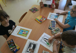 Kilkoro dzieci przy stole wykonuje pracę plastyczną z wykorzystaniem kredek i szablonów.