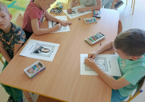 Kilkoro dzieci przy stole wykonuje pracę plastyczną z wykorzystaniem kredek i szablonów.