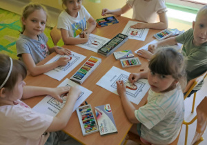 Kilkoro dzieci przy stole wykonuje pracę plastyczną z wykorzystaniem kredek i szablonów.