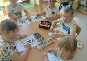 Kilkoro dzieci przy stole wykonuje pracę plastyczną z wykorzystaniem kredek i szablonów.